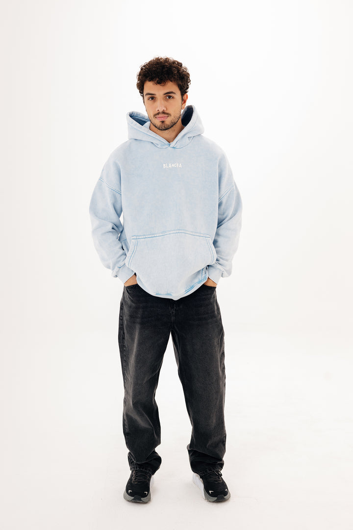 Male model wearing baby blue washed oversized hoodie with BLANCKA print — full body shot on white studio background
