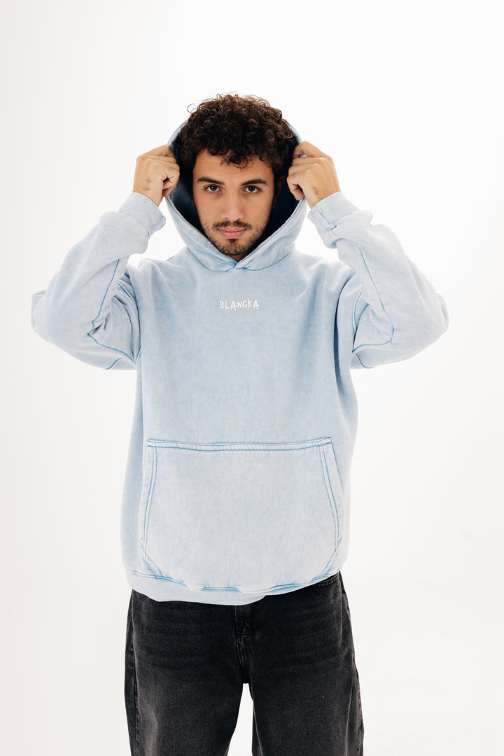 Close-up of male model in baby blue acid wash hoodie with front BLANCKA logo — studio background
