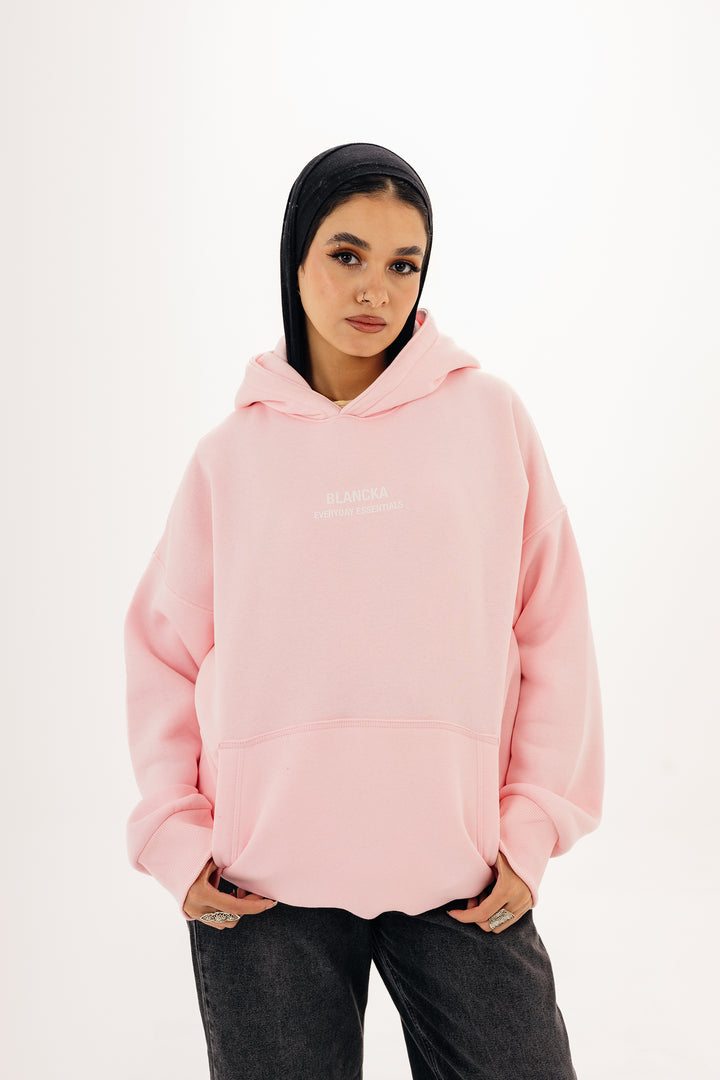 Female model wearing pink boxy hoodie with BLANCKA Everyday Essentials print — studio background
